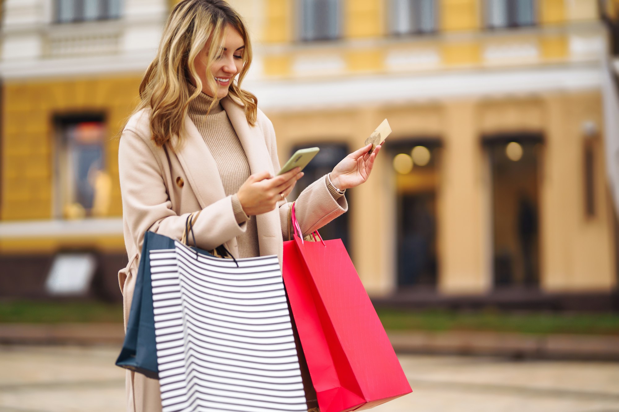 Woman with telephone and credit card. Blond girl in light coat holding shopping bags on city street